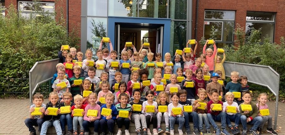 The first graders are happy about their great organic lunch boxes and proudly hold them towards the camera.