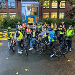 The children and teachers of Class 4a prepare to leave for the farm on their bicycles. They wear high-visibility vests and a helmet.