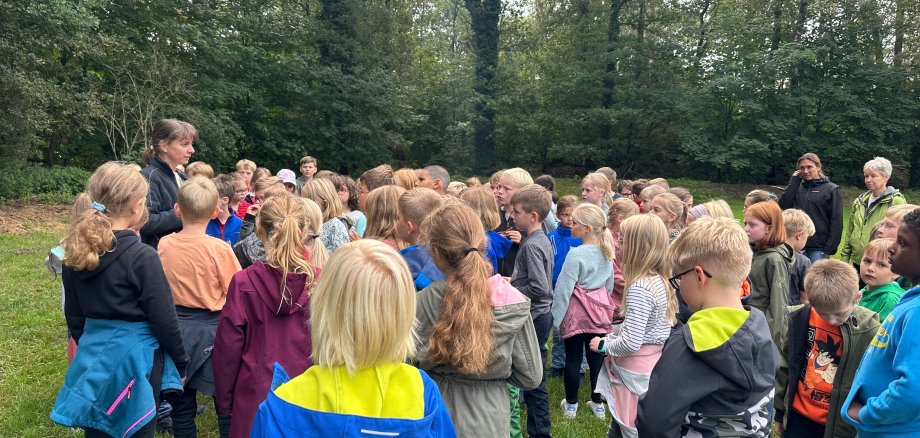 Een leerkracht staat op een weiland en geeft uitleg terwijl de kinderen aandachtig luisteren. Op de achtergrond is het bos te zien.